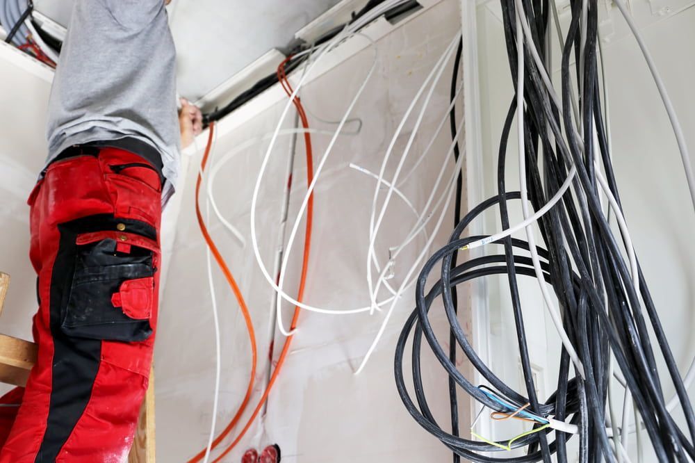 A Man Is Working On A Wall With A Bunch Of Wires Hanging From It — Greg Keft Electrical in Port Macquarie, NSW