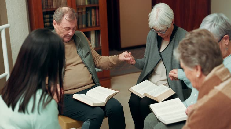 A group of diverse people, seated in a circle, praying with their heads bowed, holding open books.