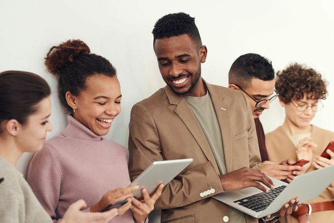 Group of young adults smiling at tablets, laptop, and phones near a white wall.