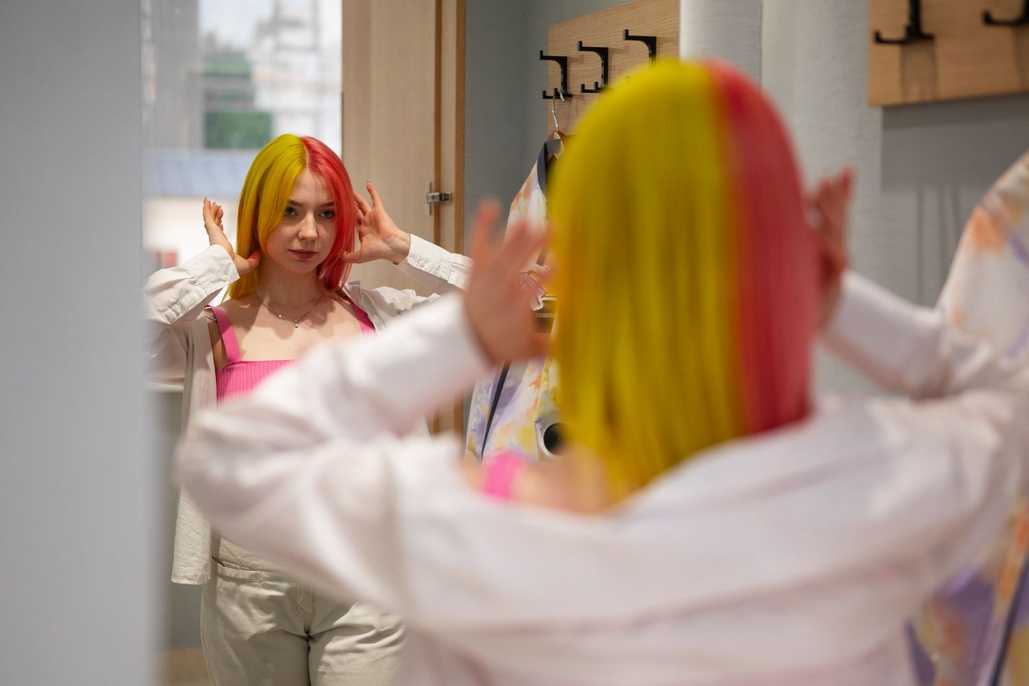 A person with half-yellow, half-pink hair looks into a mirror while adjusting their hair in a room with wall hooks.