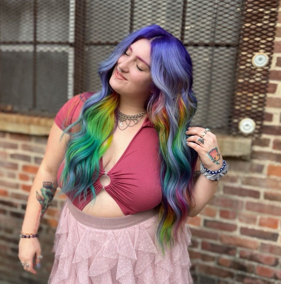 A woman with rainbow hair is standing in front of a brick wall.