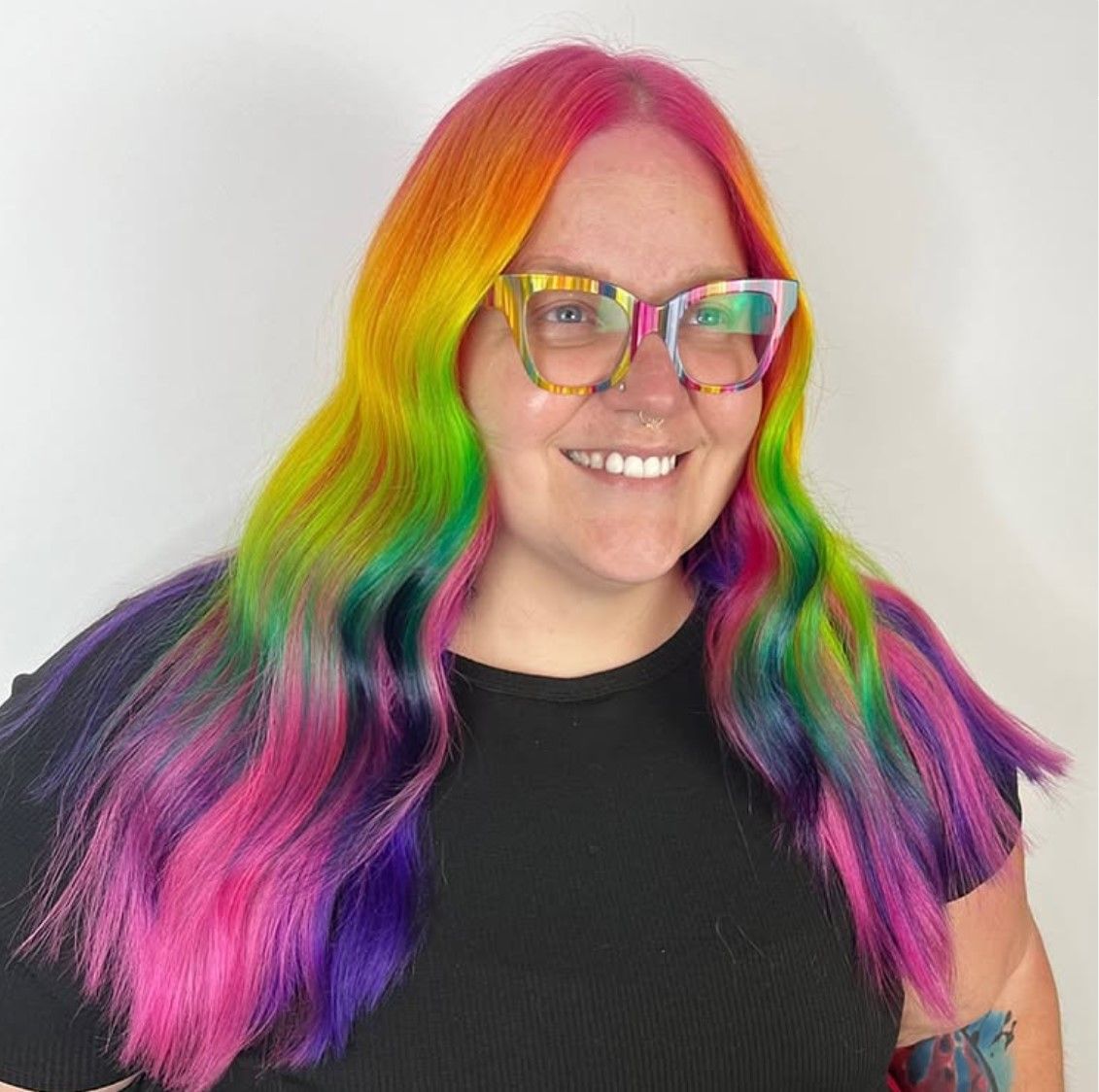 A woman with rainbow hair and glasses is wearing a black shirt.