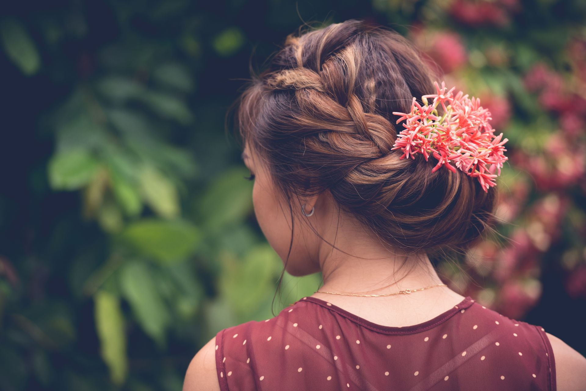 Woman with braided updo, adorned with pink flowers, in a maroon dress; outdoors.