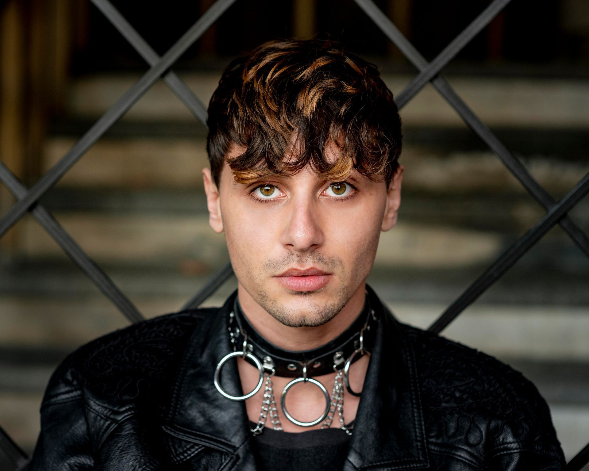 A young man wearing a choker and a leather jacket is sitting in front of a fence.