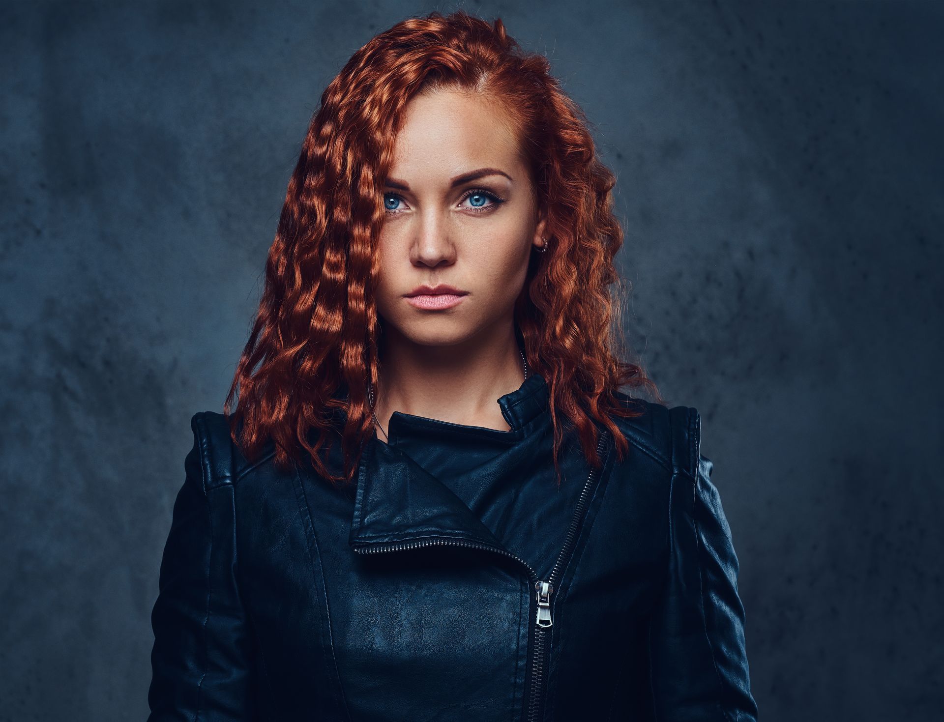 Woman with red curly hair in a black leather jacket, looking forward against a gray background.