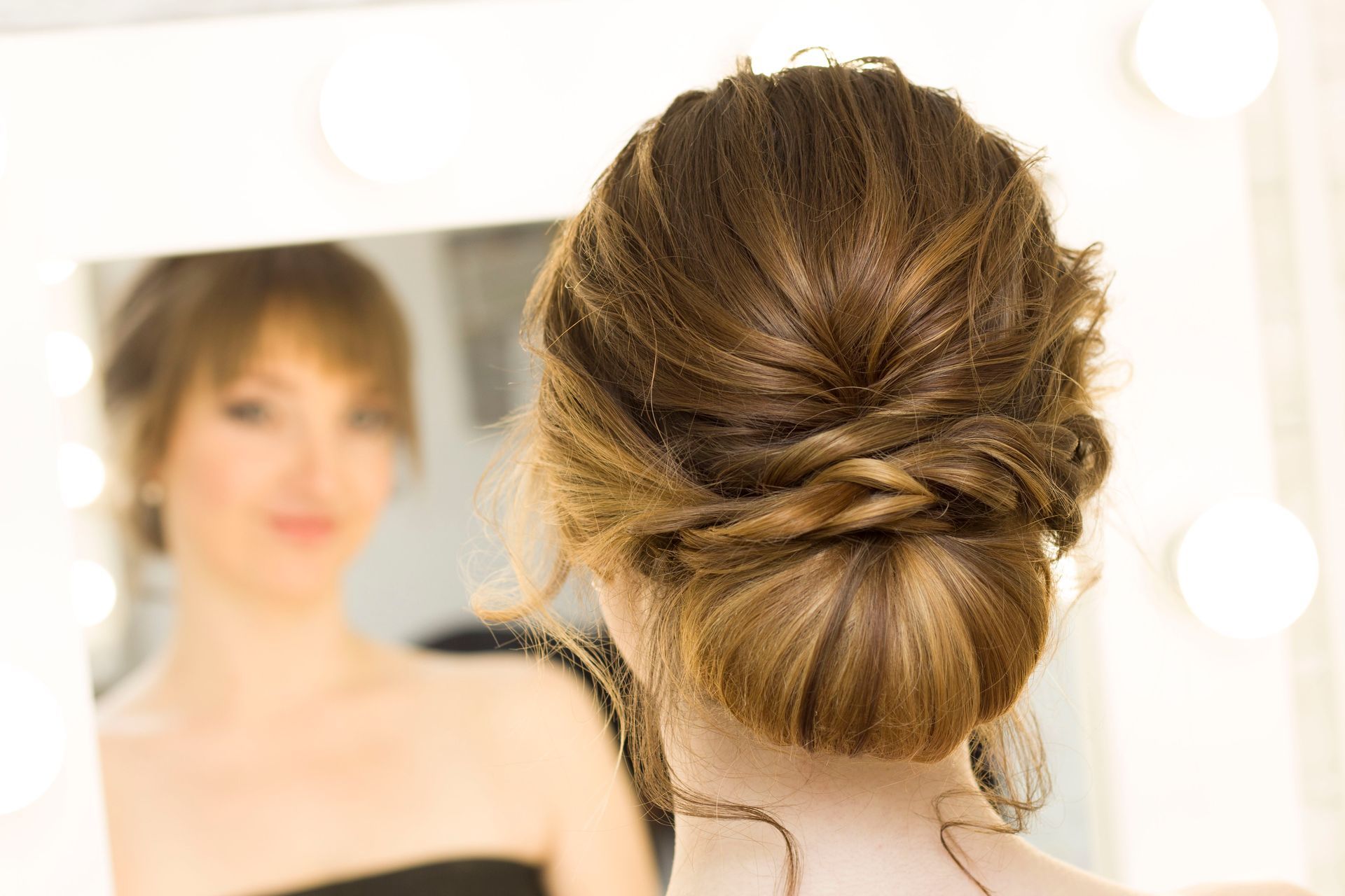 Woman with brown hair styled in an updo, looking into a lit mirror.