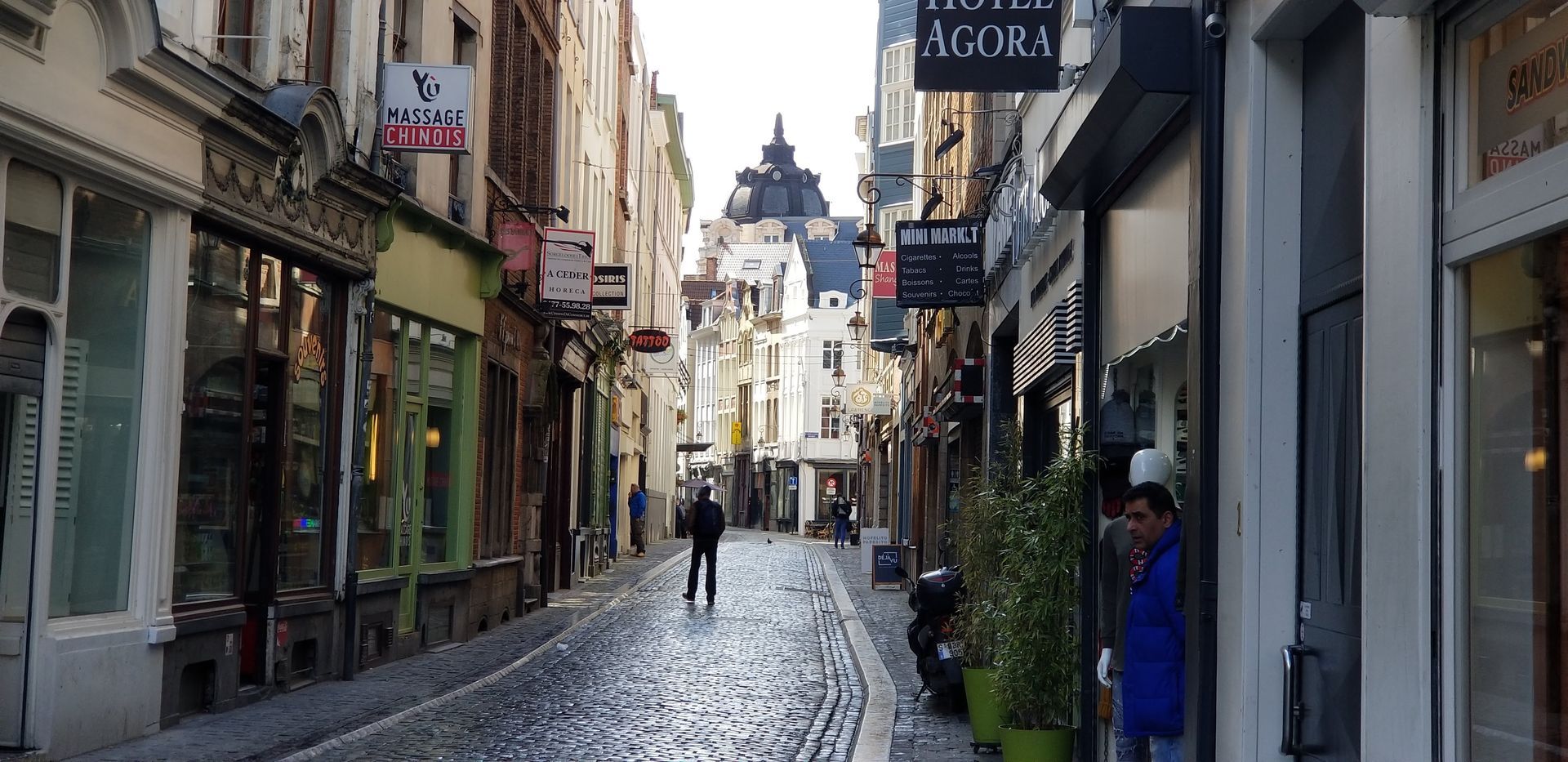 Narrow European cobblestone street with shops and a hotel sign. A person walks down the street.
