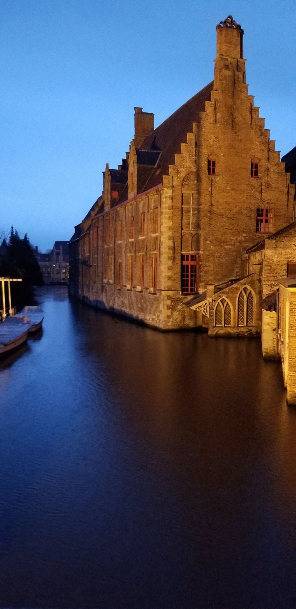 Canal in Bruges, Belgium, with a large brick building on the right under a twilight sky.