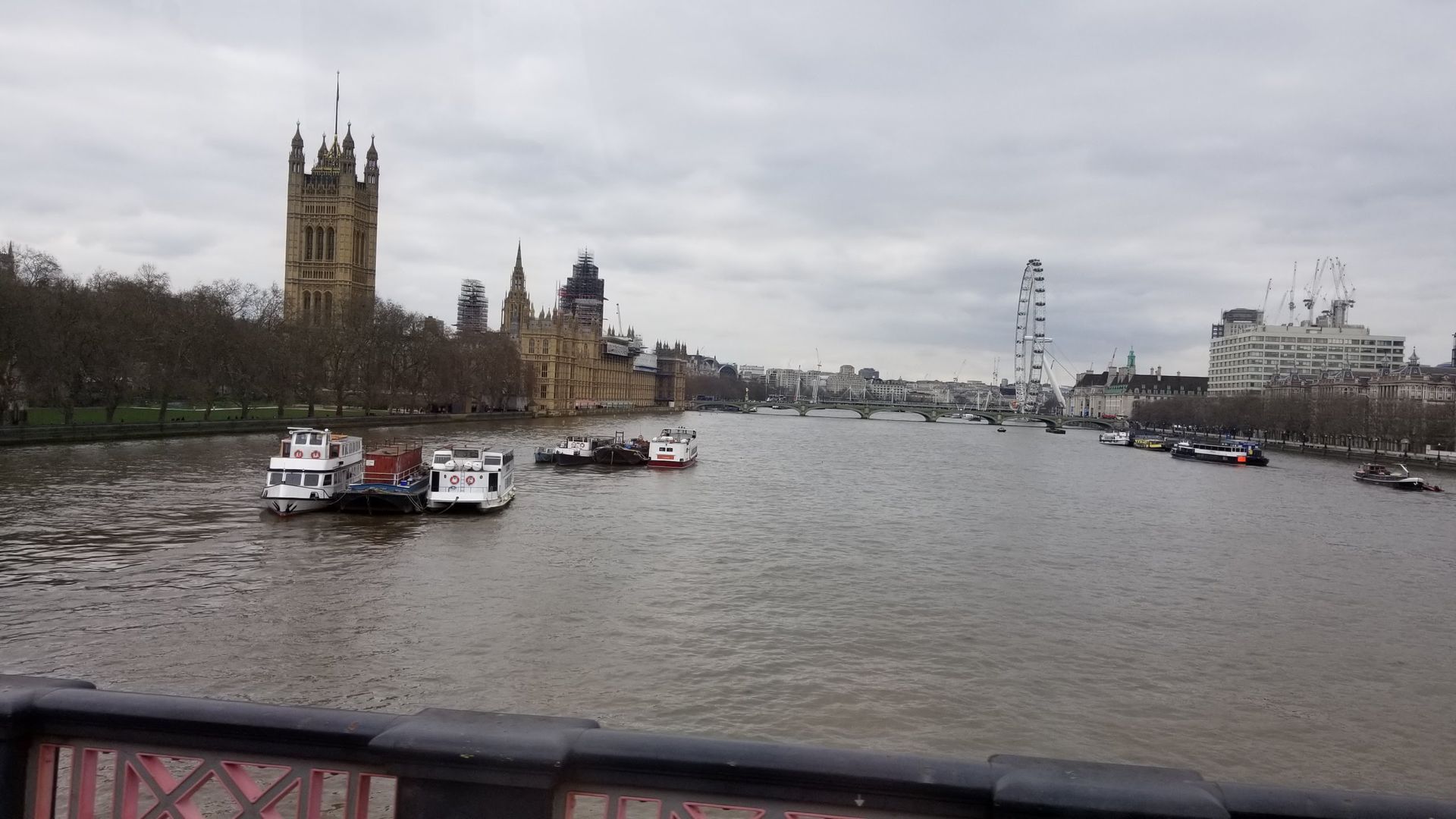Thames River view of the Houses of Parliament, London Eye, and boats on a cloudy day.