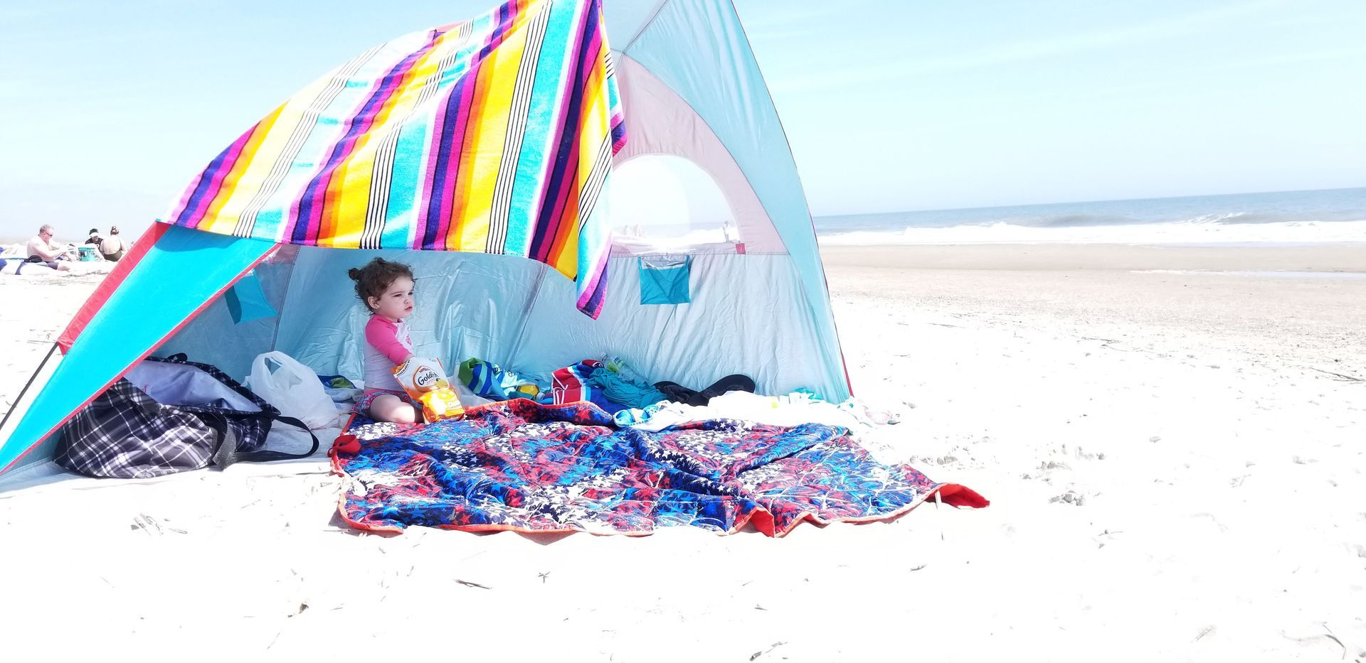 Child under a beach tent on a sunny beach, waves in the background.