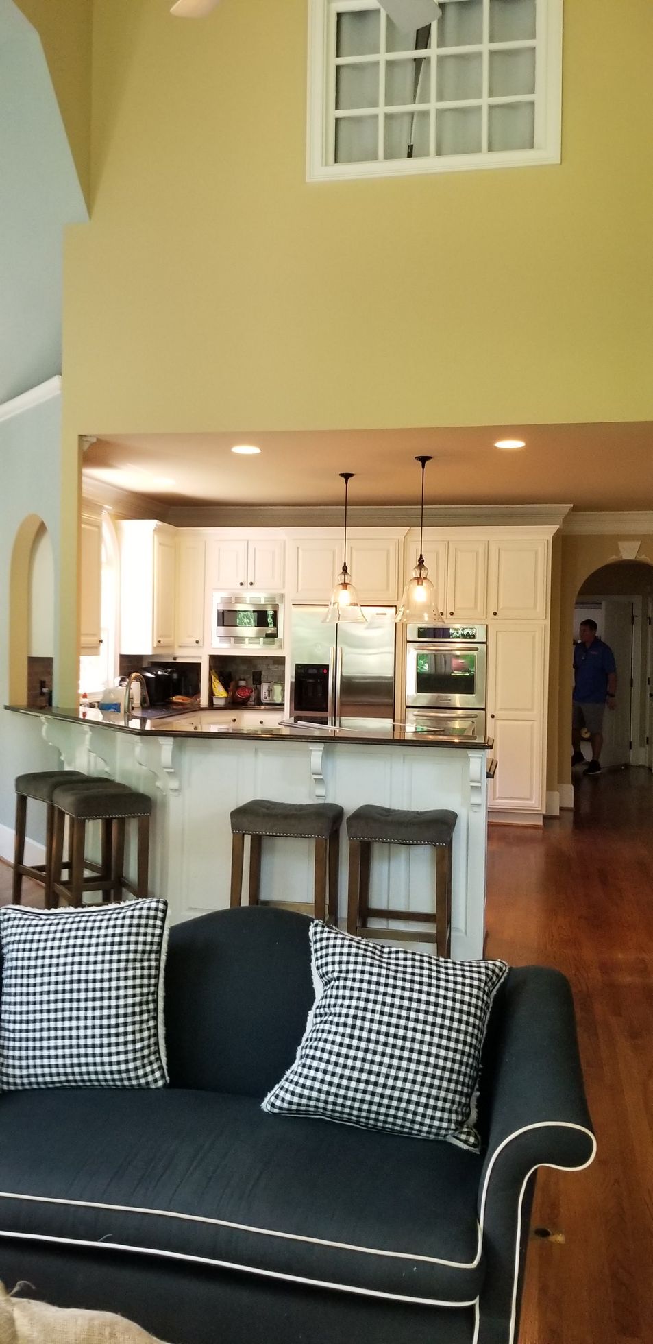 Living room with open kitchen, black sofa, checkered pillows, stools, and pendant lights.