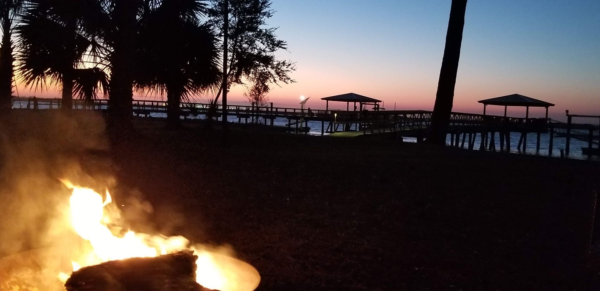 A bonfire burns on a beach as the sun sets over docks and a calm sea.