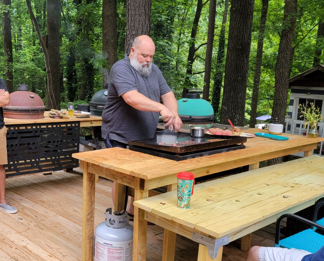 Man grilling on a flattop griddle outdoors. He's near a table and bench on a deck with trees in the background.