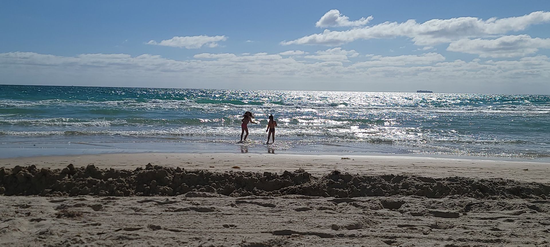Two people walk on a beach near the water. The sun reflects on the waves under a blue sky.