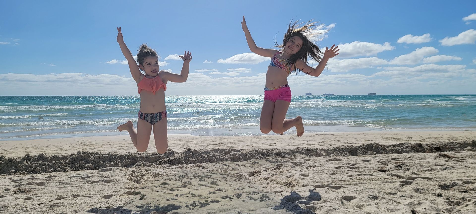 Two girls in swimwear jump on a sandy beach near the ocean, arms raised. Bright blue sky with clouds in background.