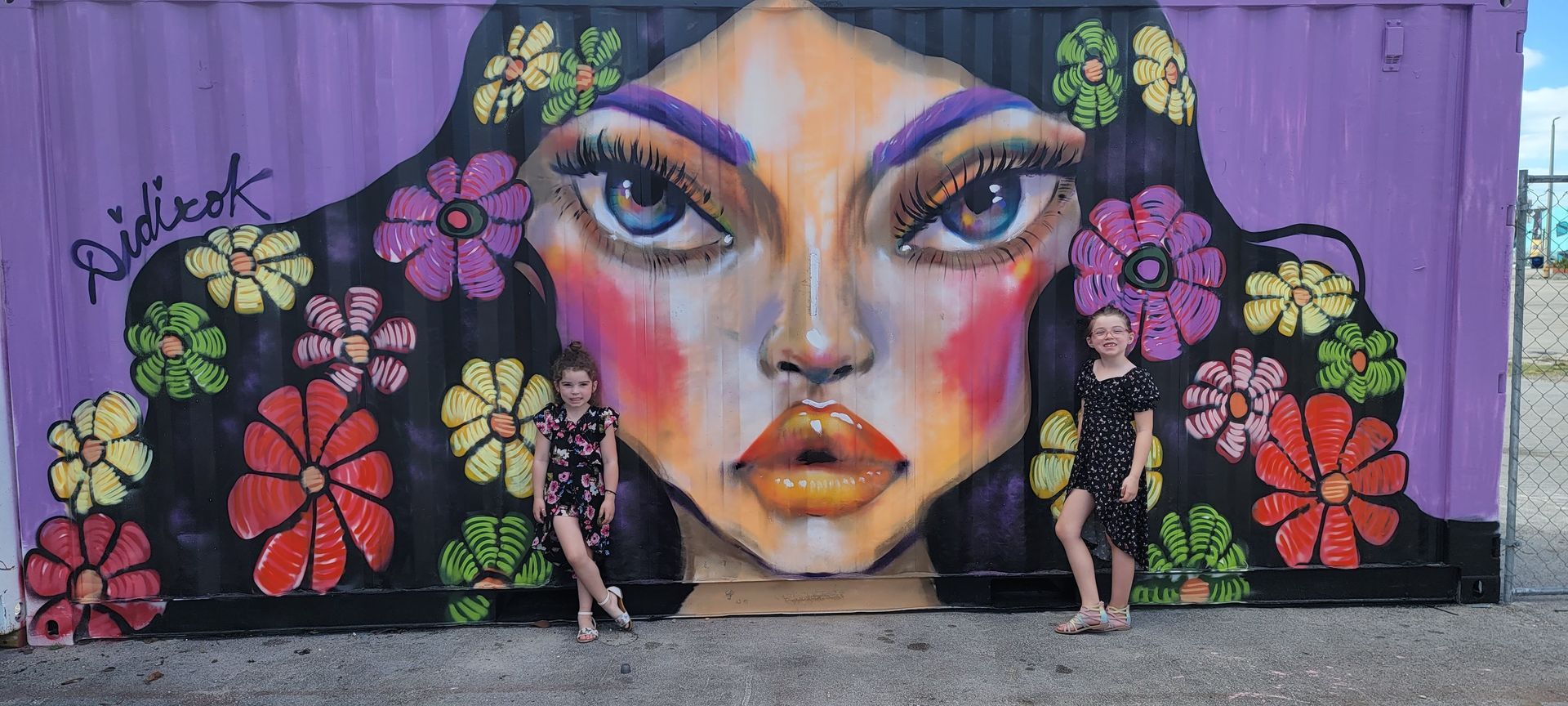 Two people stand in front of a colorful mural of a woman's face with flowers in her hair.