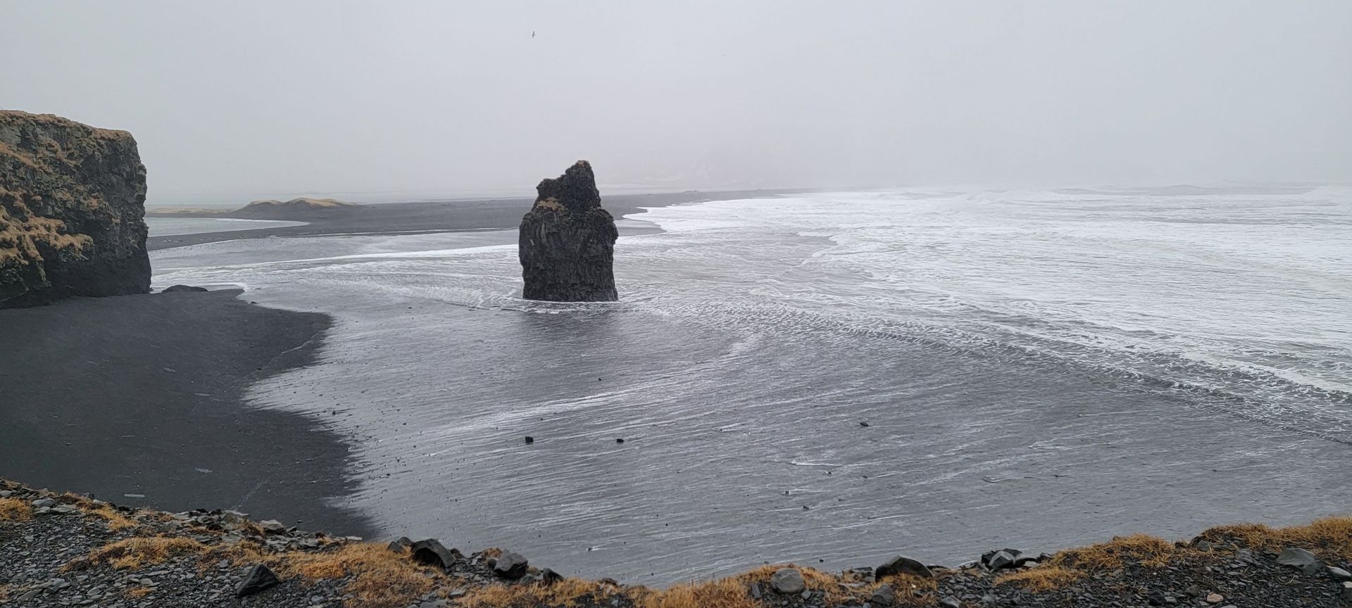 Black sand beach with a large rock formation in the sea and a cliff to the left, overcast sky.