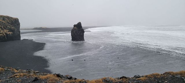 Black sand beach with a large rock formation in the sea and a cliff to the left, overcast sky.