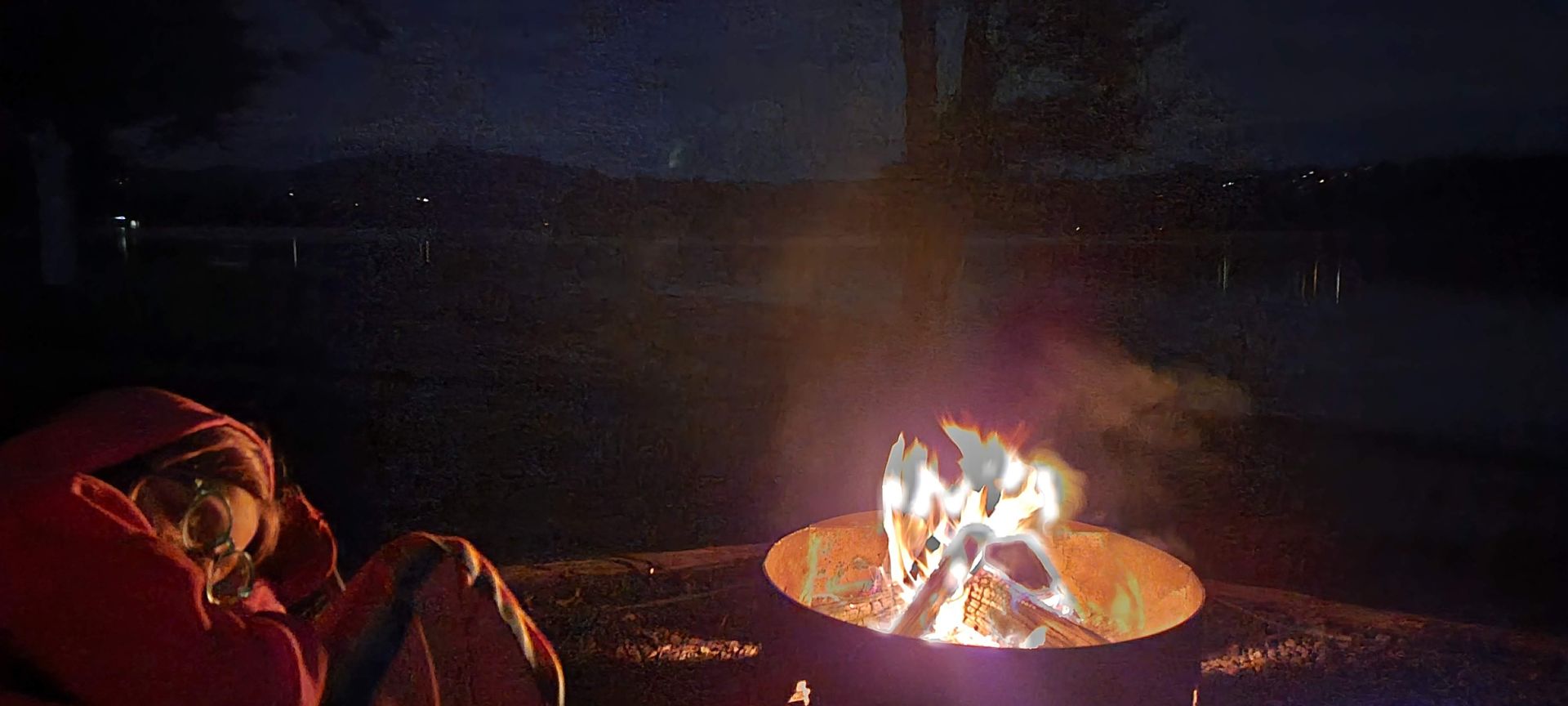 Person by a campfire at night overlooking a lake. The fire glows orange.