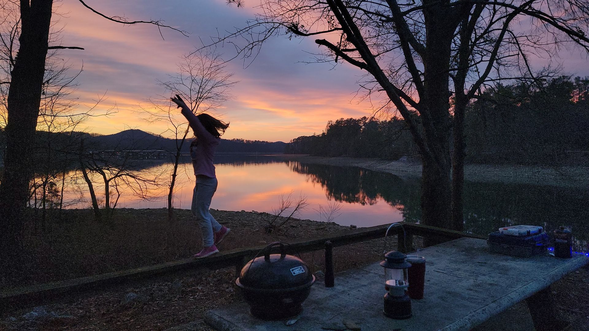 Person balances on a railing, arms raised, by a lake at sunset. Trees and a small grill are also visible.
