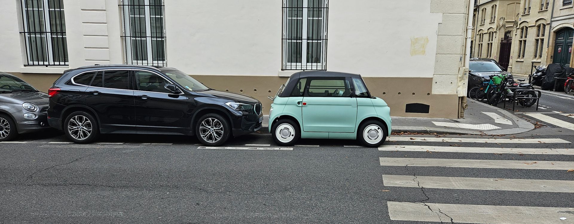 Cars parked on a street near a crosswalk. One is a small blue car, next to a black SUV.