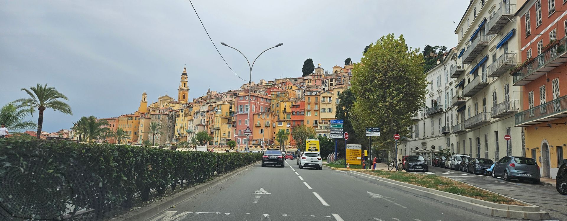 Road leads to colorful buildings on a hill, a coastal town. Cars, hedges, and cloudy sky.