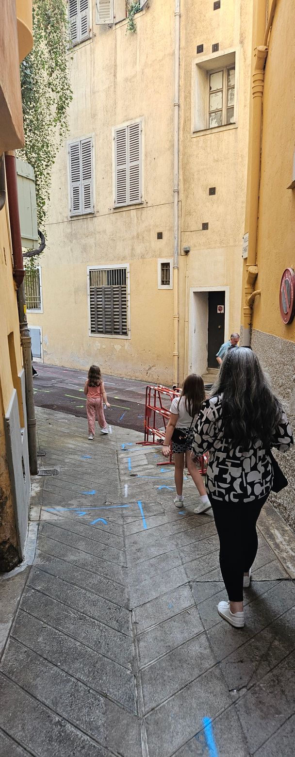 People walking up a narrow, stone street between old buildings with shuttered windows.