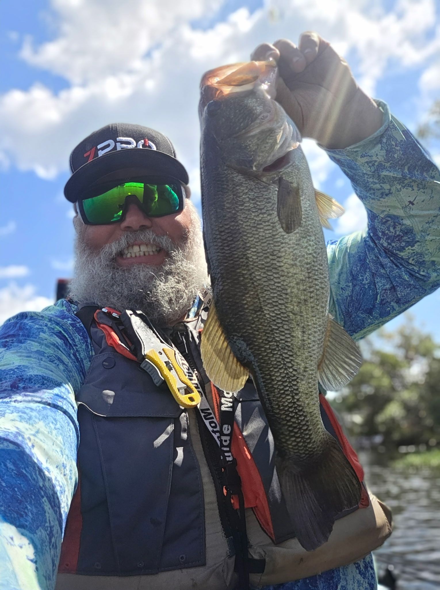 Man holding up a large bass. He wears sunglasses, a cap, and a fishing shirt. Blue sky in the background.