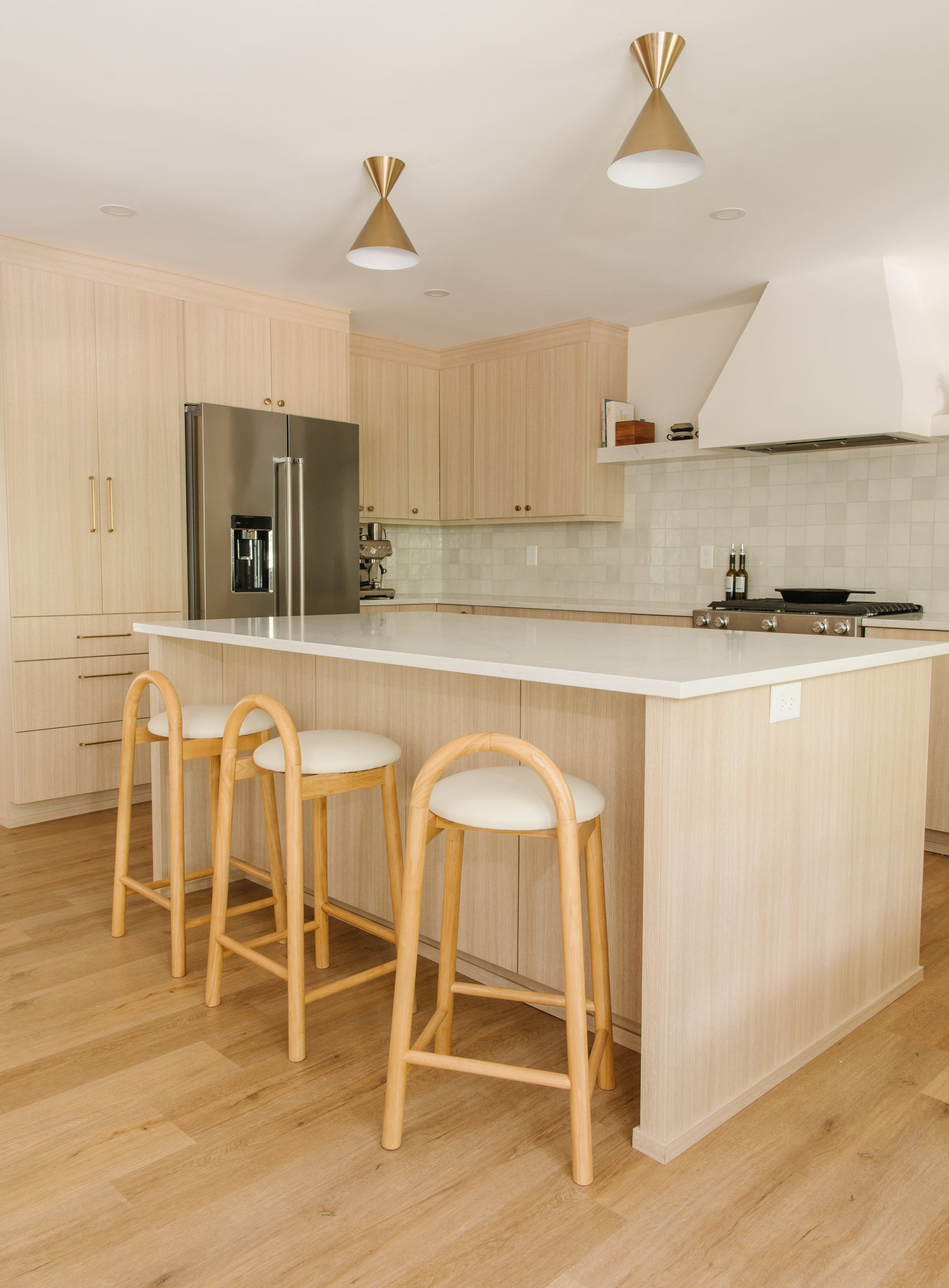 Kitchen with light wood cabinets, island, and stools, stainless steel refrigerator, and gold pendant lights.