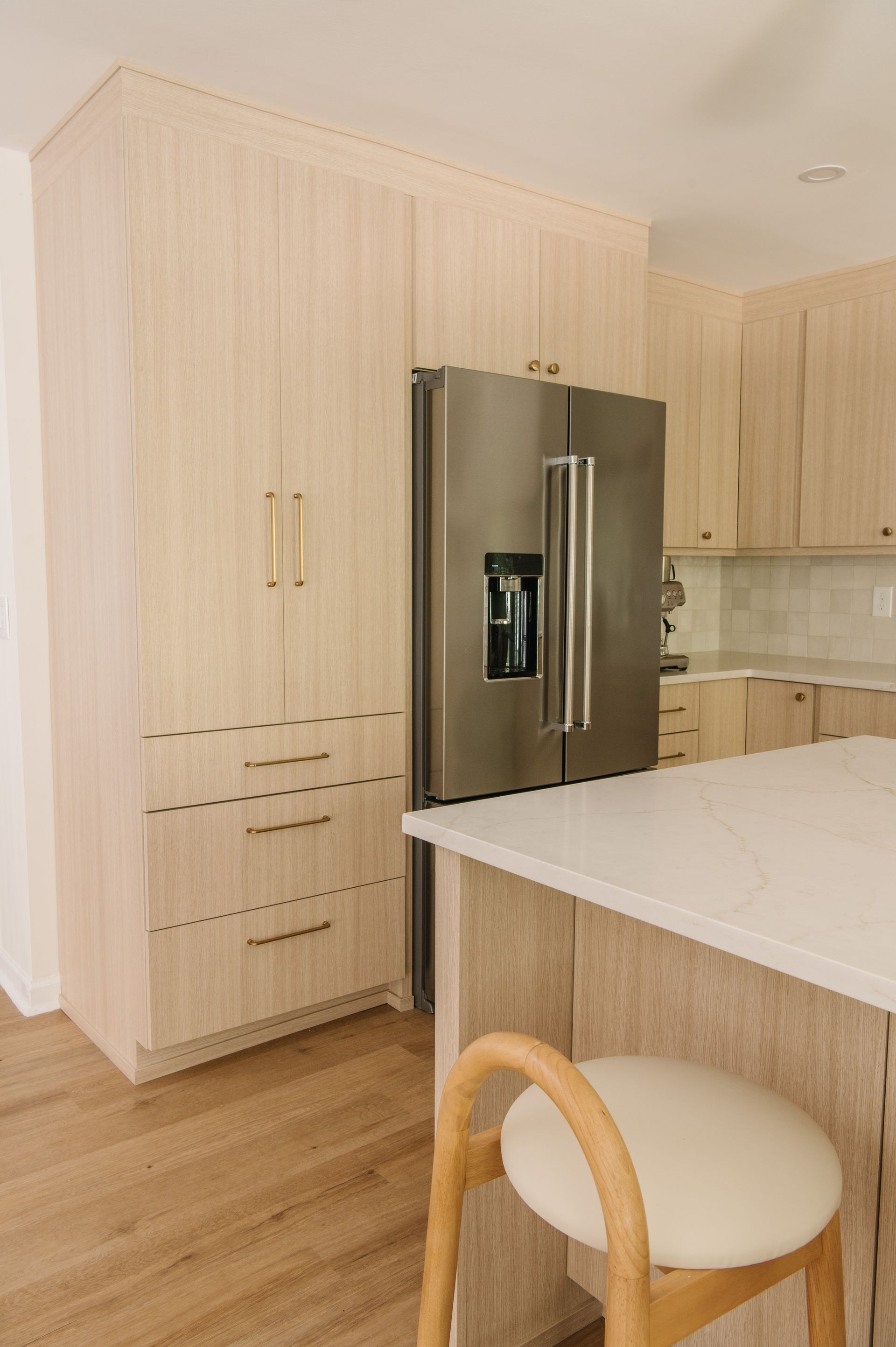 Light-toned wood kitchen with stainless steel refrigerator and white countertop island. Stool in foreground.