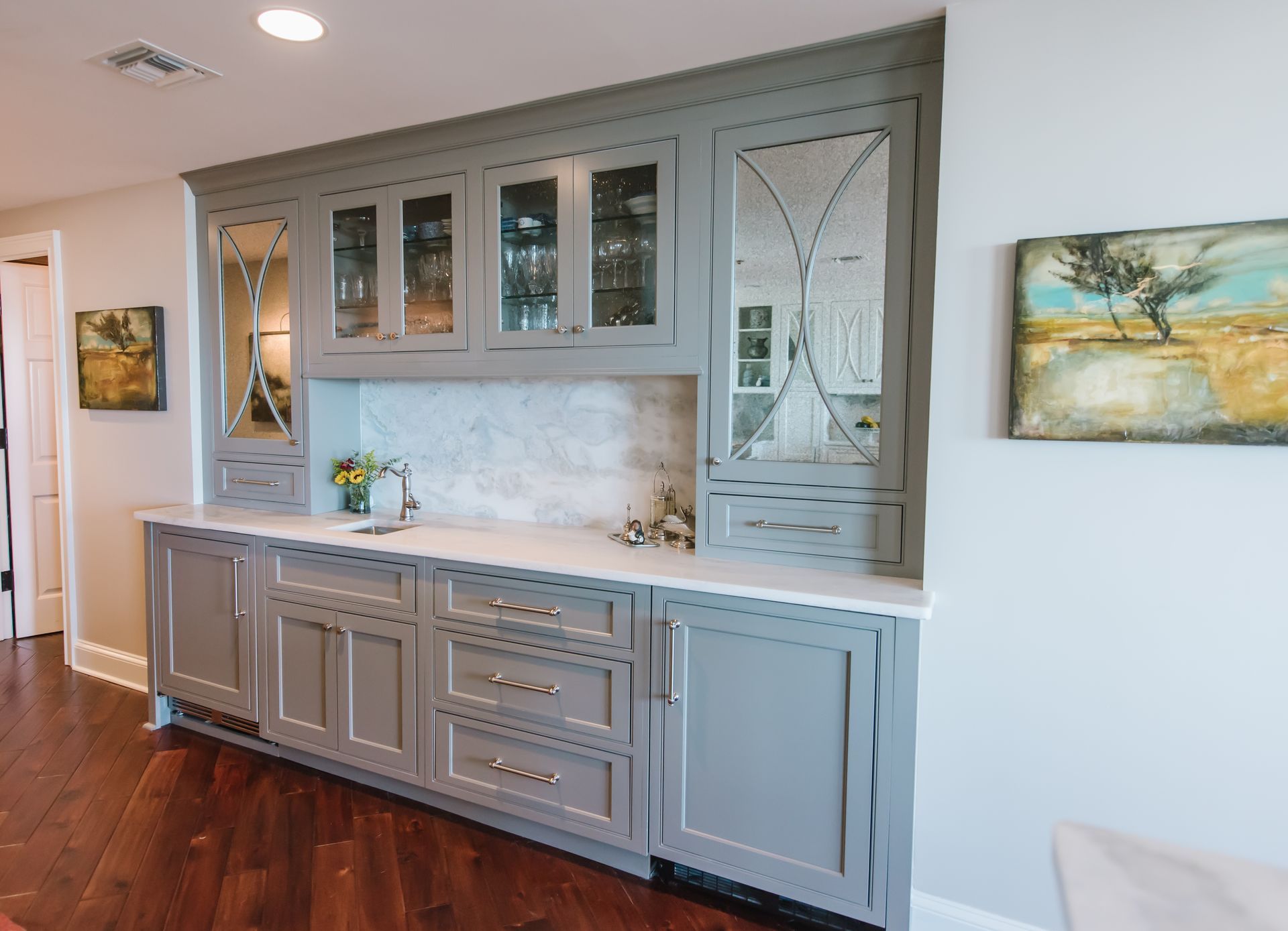 Gray built-in cabinet with mirrored panels and white countertop against a white wall; art on the wall and wooden floor.