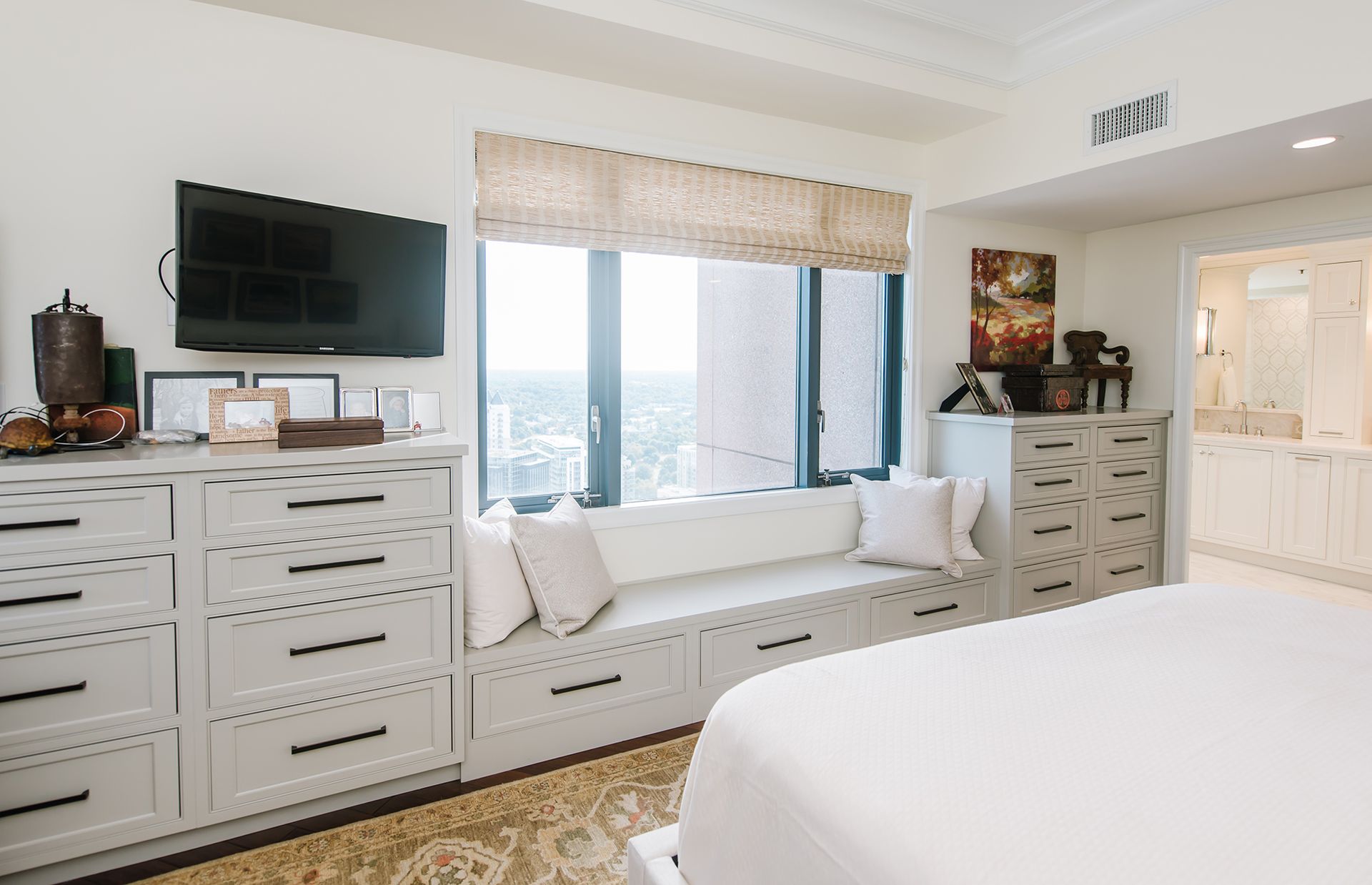 Bright bedroom with window seat, dressers, television, and view of city skyline.