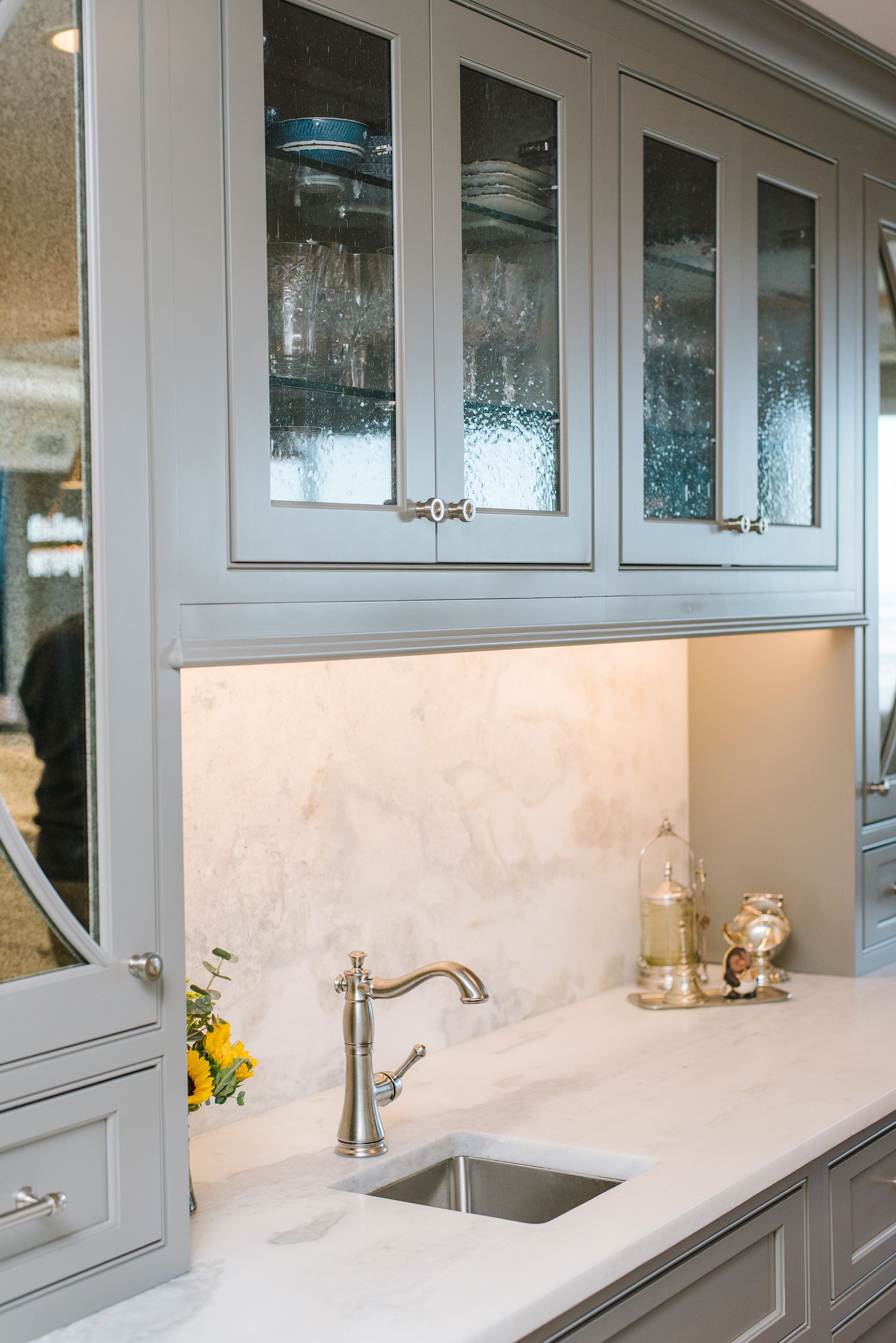 Gray kitchen cabinets with glass doors over a sink, lit by under-cabinet lights.