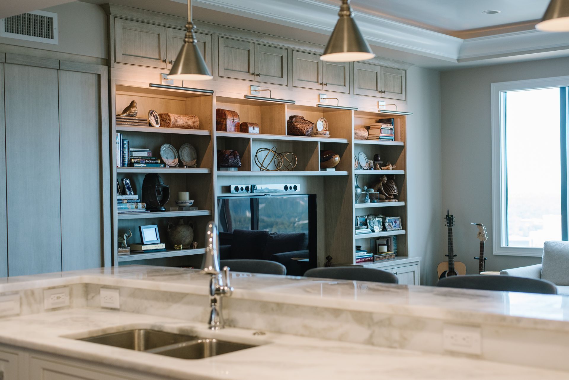 Kitchen with built-in shelves, TV, and marble countertop. Two pendant lights hang over a double sink.