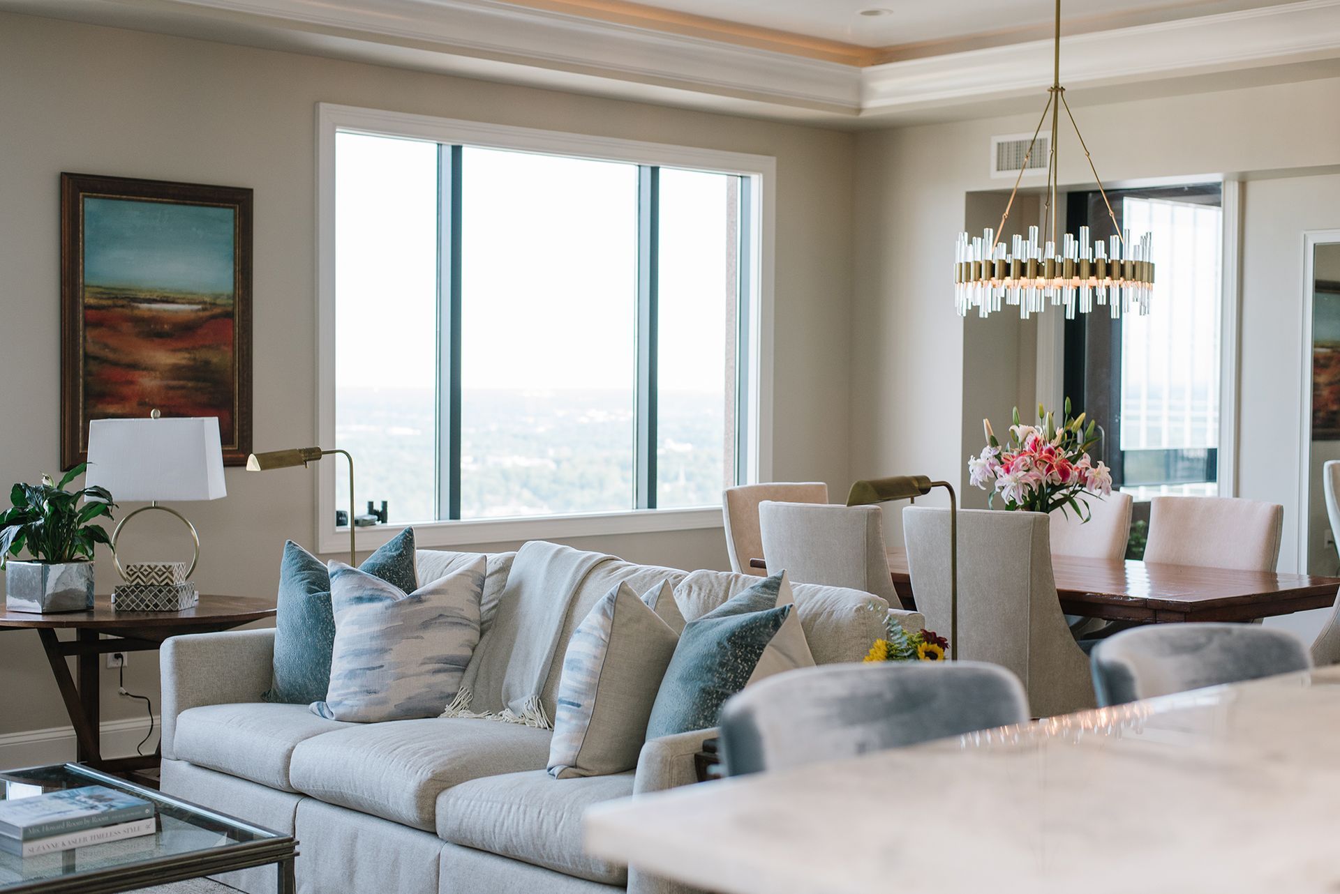 Living room with sofa, dining area, and large window overlooking a cityscape; soft colors, elegant design.