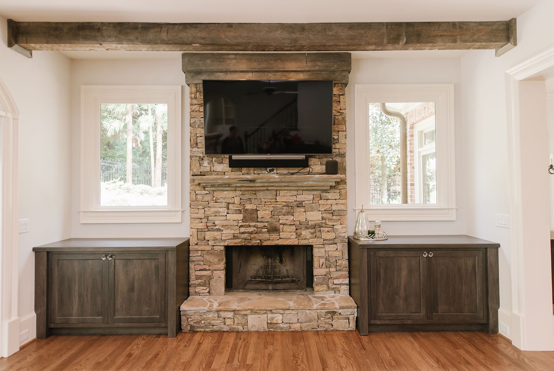 Stone fireplace with TV and windows, flanked by gray cabinets in a room with wood floors.