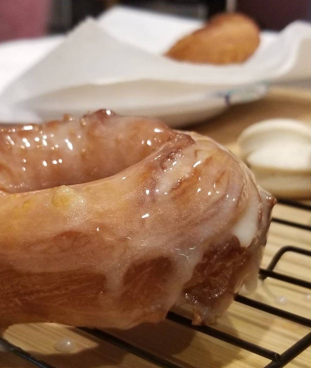 Glazed donut on a cooling rack, with a blurred background showing another pastry and plate.