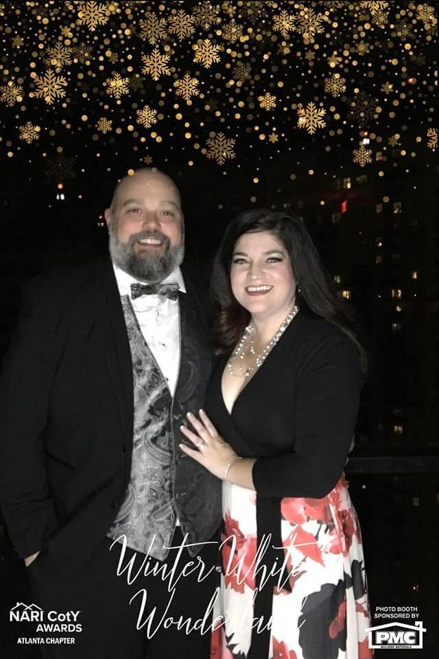 Couple posing in front of a backdrop with gold snowflakes. Man in tuxedo, woman in black top and floral skirt.