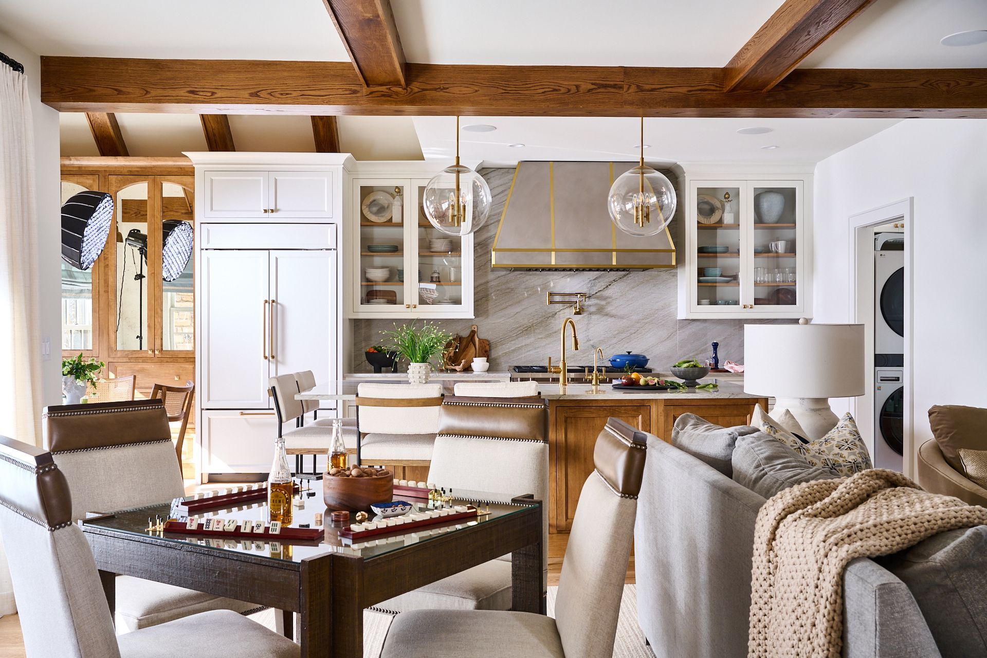 Kitchen with a dining table in front.  White cabinets, marble backsplash, and wooden beams.