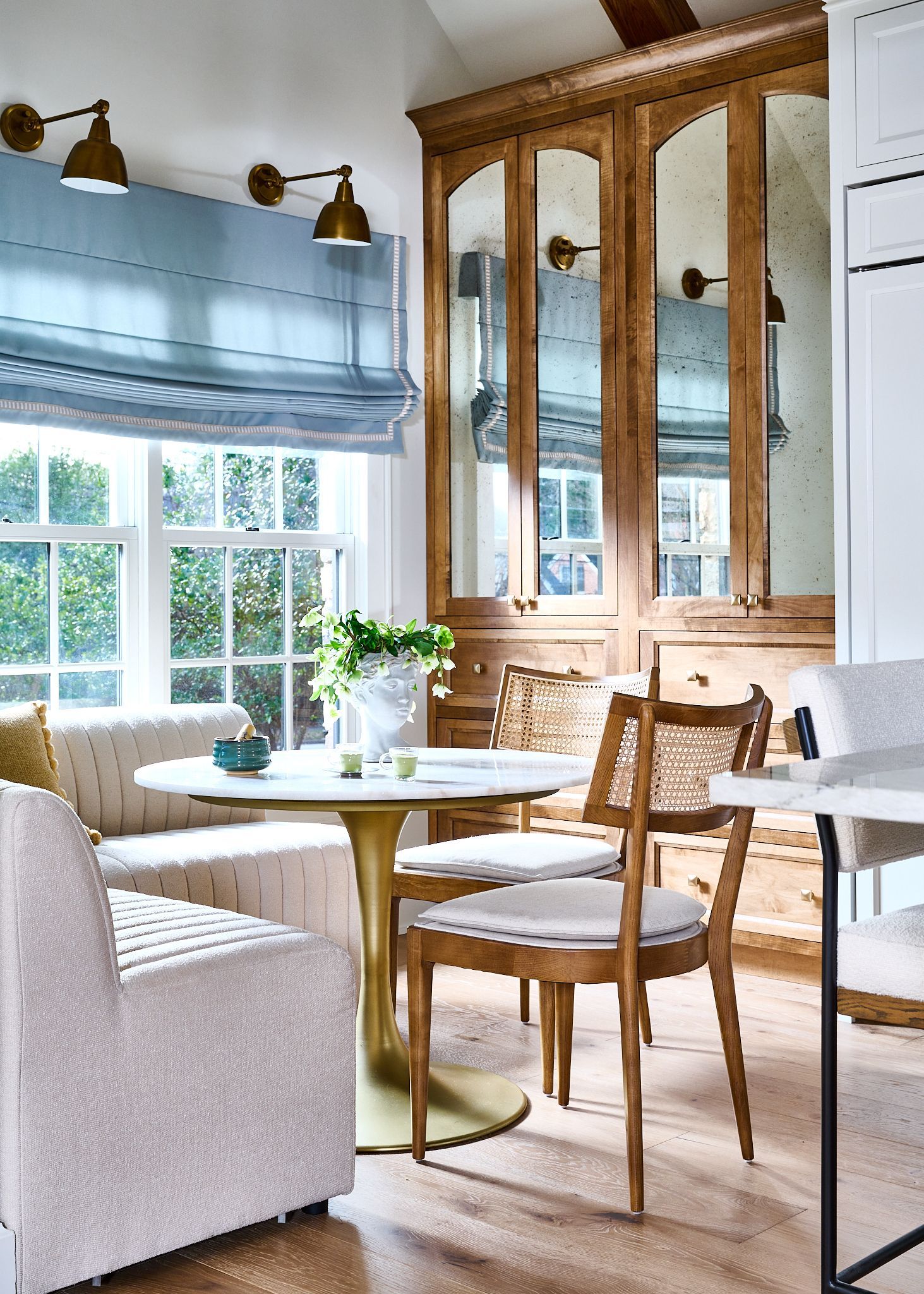 Cozy breakfast nook with light blue roman shade, round table, gold accents, and vintage mirrored cabinet.