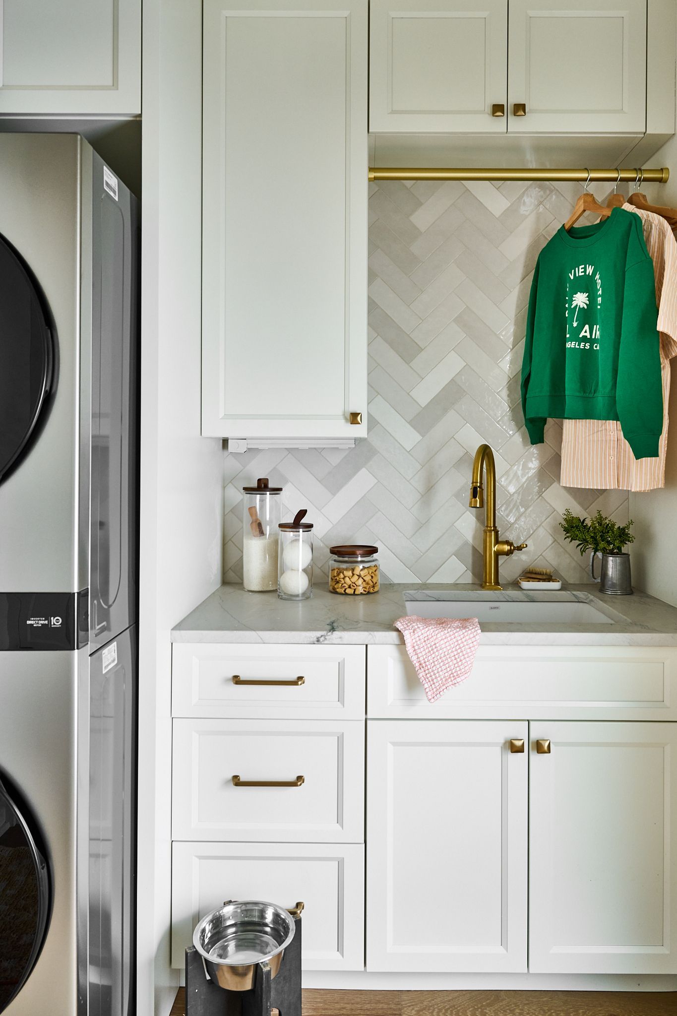 Laundry room with stacked washer/dryer, sink, cabinets, and hanging clothes. White and gold accents.