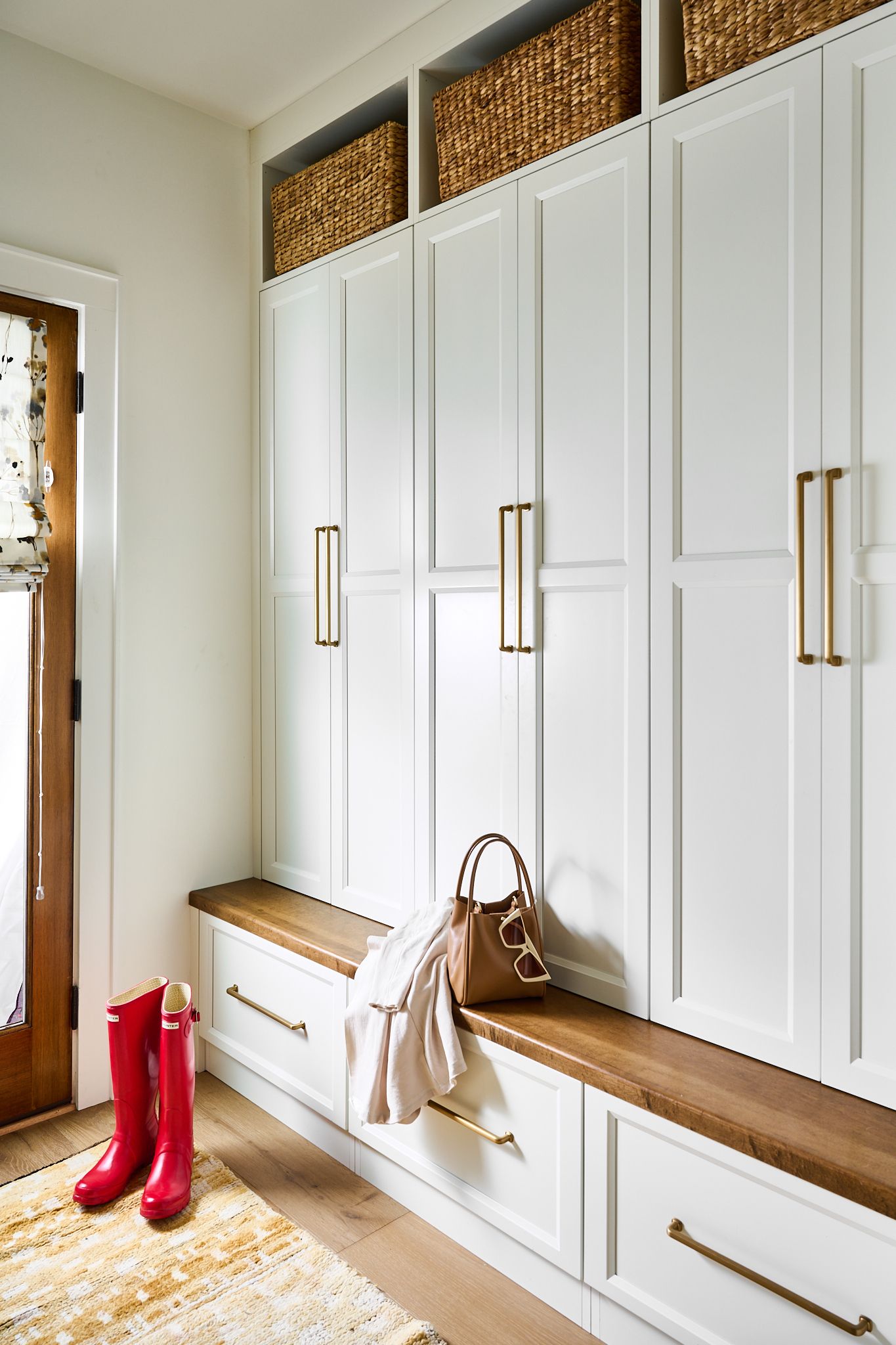 White mudroom with bench, storage cabinets, baskets, wood accents, red boots, and handbag.