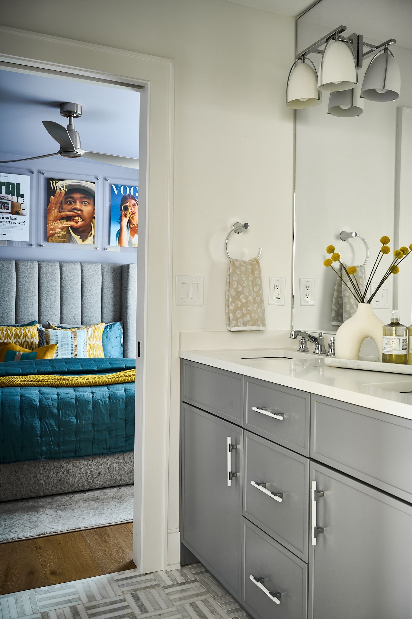 Bathroom with gray vanity, open doorway to bedroom with turquoise bed and framed posters.