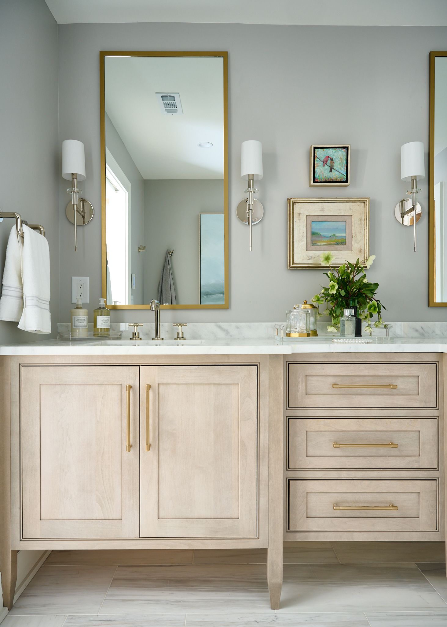 Bathroom with light wood vanity, gold framed mirror, wall sconces, and artwork. Gray walls.