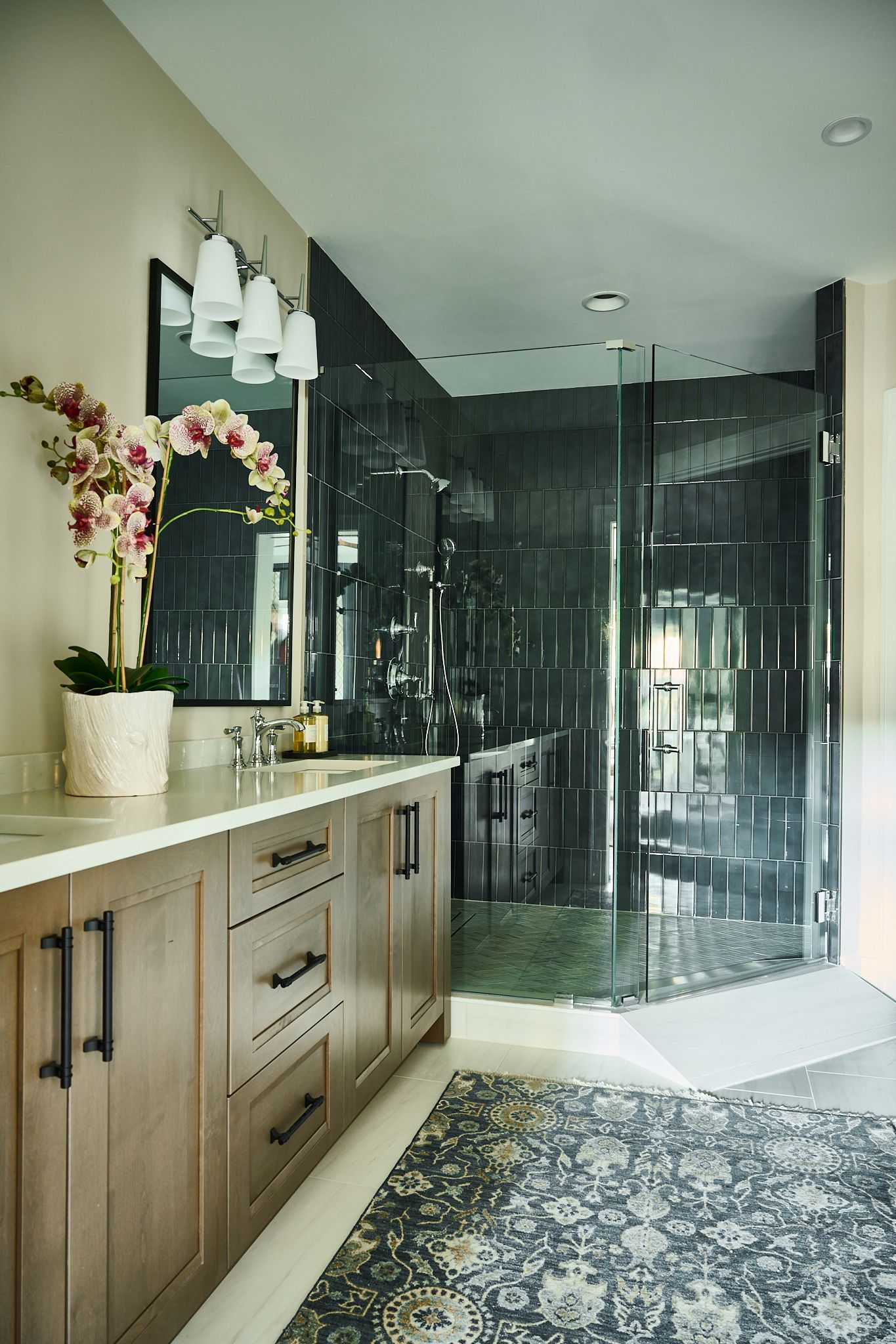 Bathroom with wooden vanity, patterned rug, glass shower, and orchid plant.