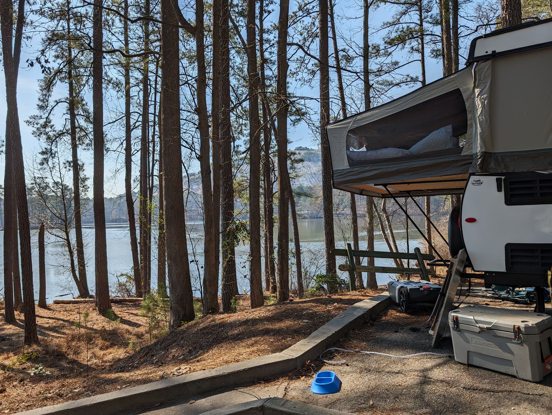 Campsite with trailer. Trees and a lake in the background. Trailer's top is open for sleeping.