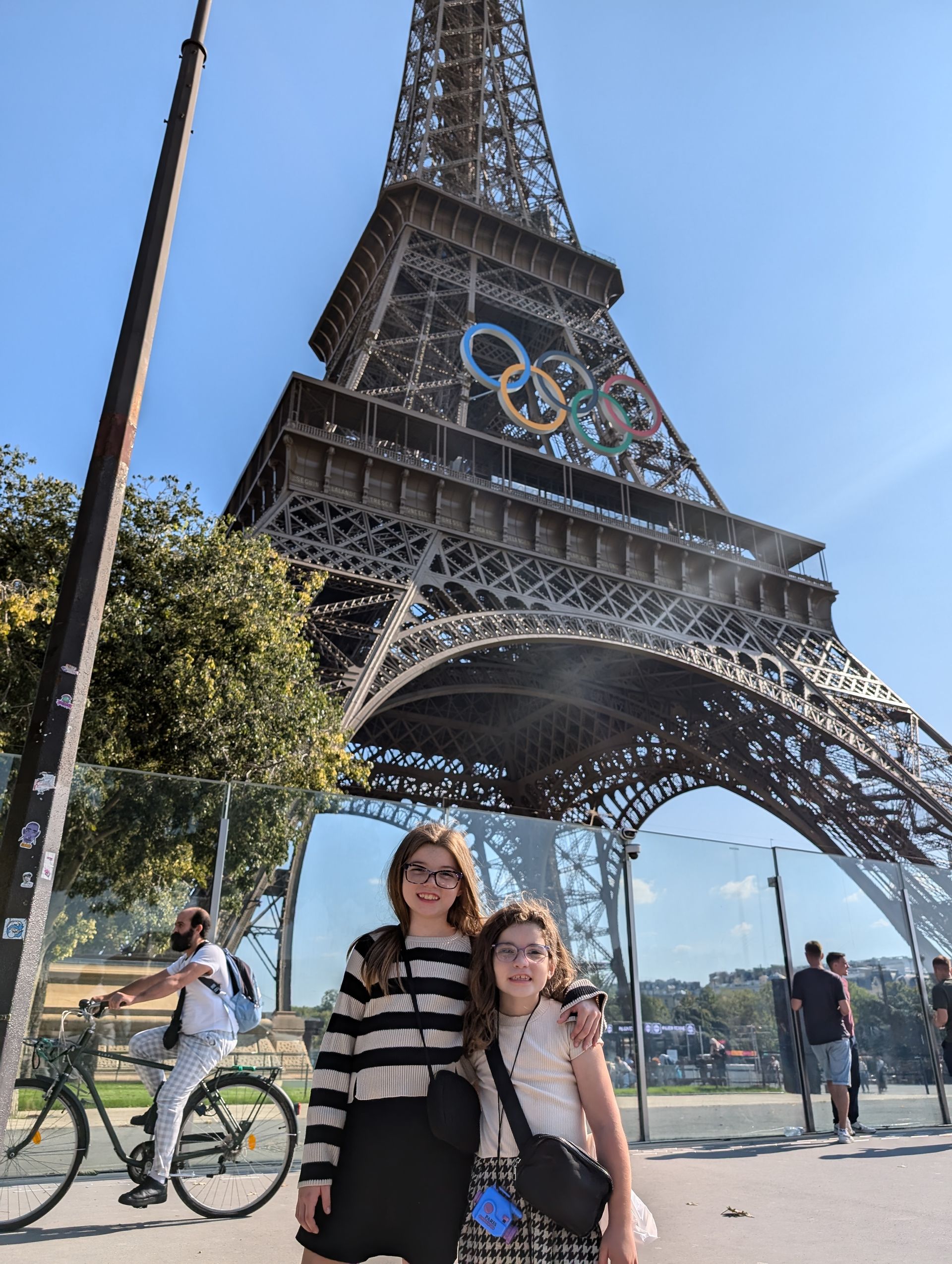 Two girls pose in front of the Eiffel Tower, adorned with Olympic rings. Sunny day.