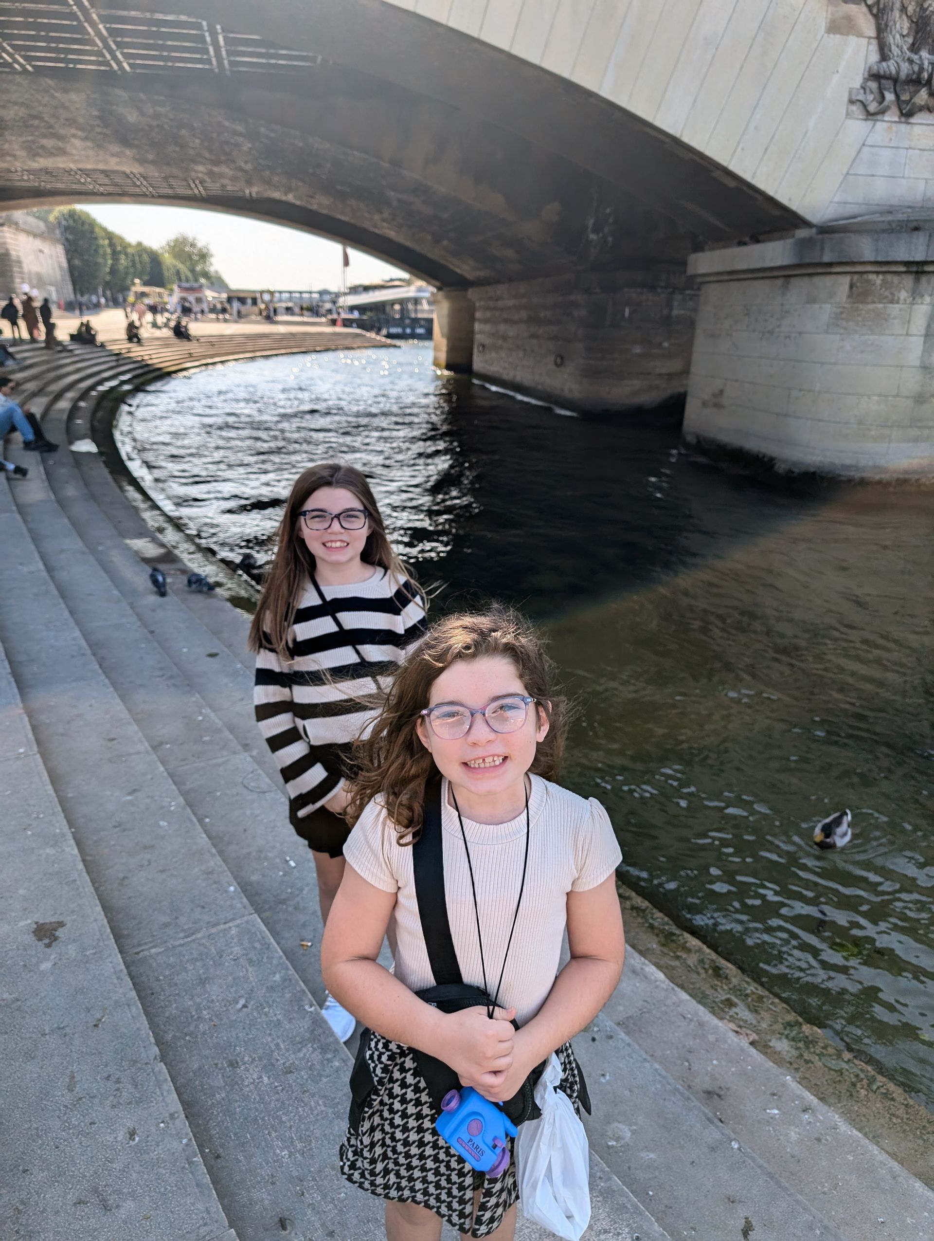 Two girls by a river in Paris, under a bridge. One in striped top, both wearing glasses.