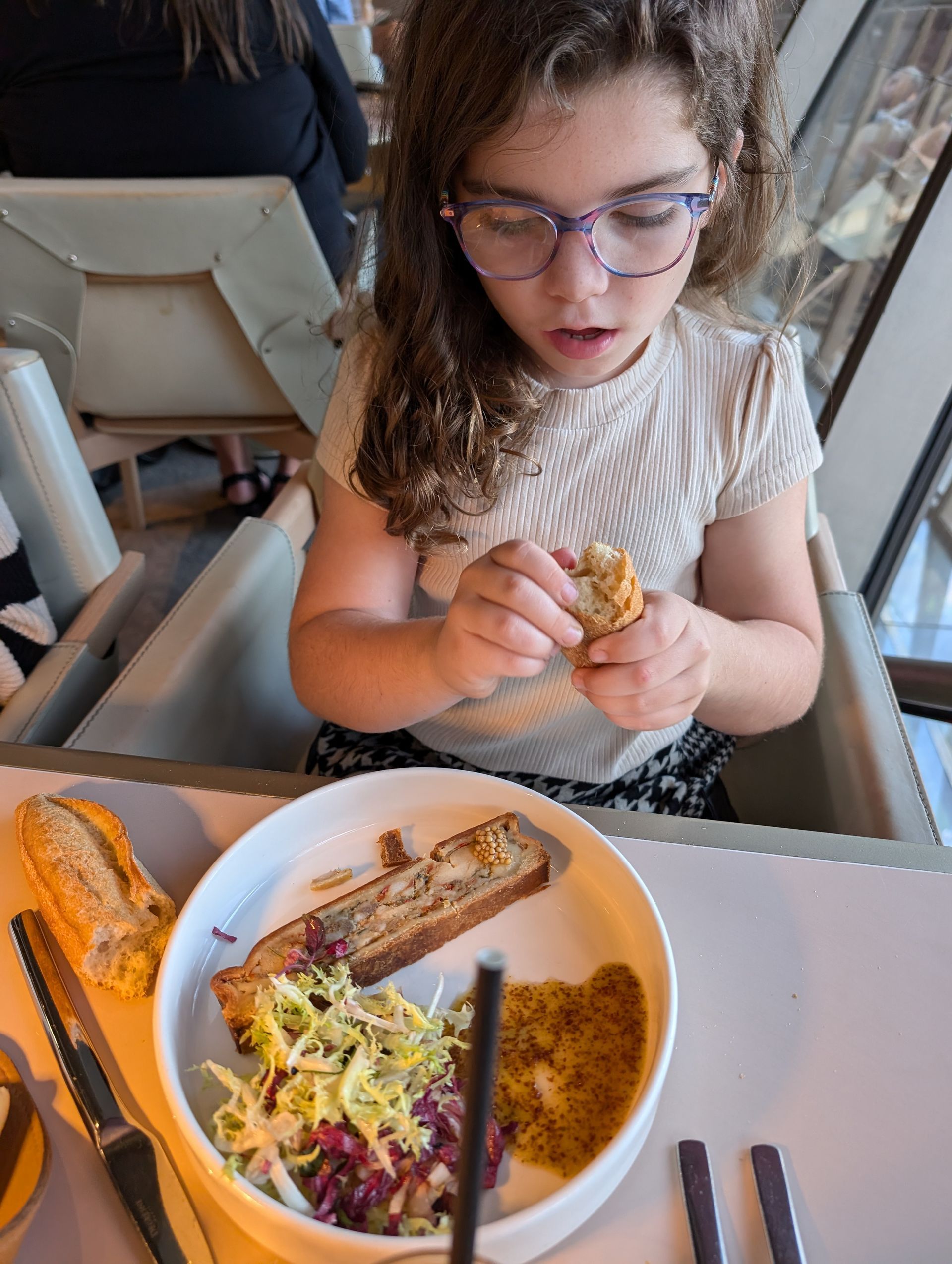 Young person in glasses looks at food in a white bowl with bread, slaw and a yellow item, eating a piece.