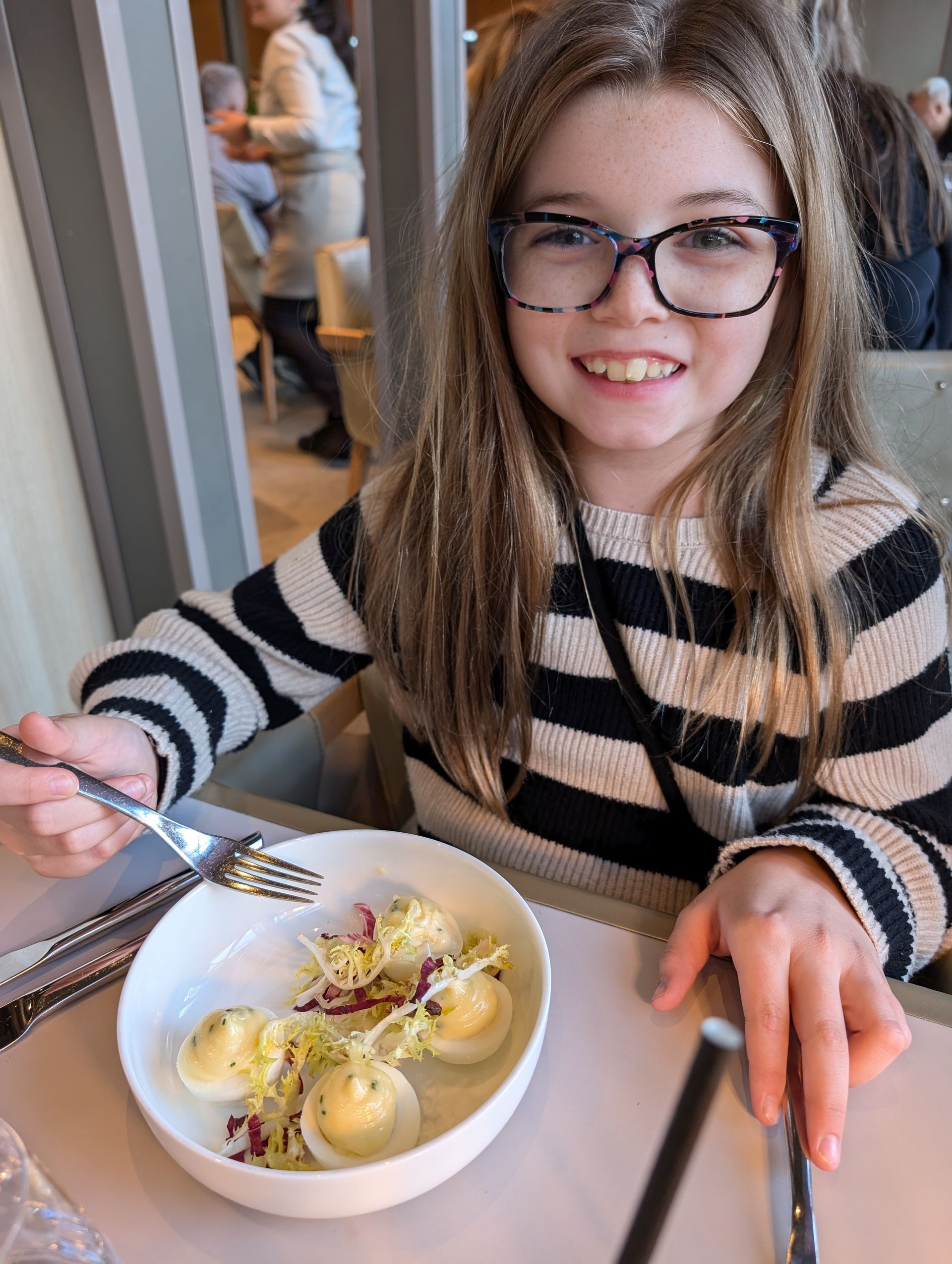 Girl with glasses smiles at the camera, eating deviled eggs in a restaurant.